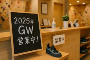 室内クライミングジムの受付カウンターに置かれた黒板に「2025年GW 営業中！」と書かれた案内。登山シューズや消毒液も置かれており、清潔感と営業中の安心感が伝わる。